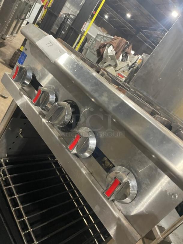 Cookline stainless steel 4 burner range/oven combo with backsplash, overhead salamander shelf, wired racks, and gas hose on casters.