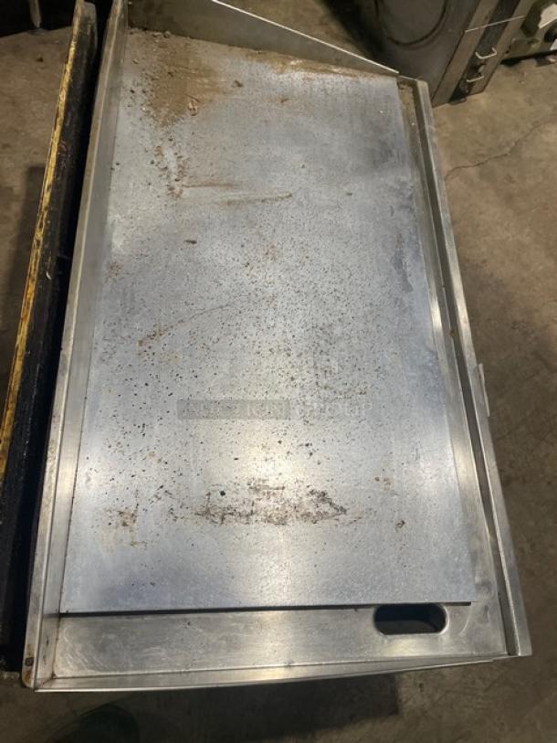 Stainless steel countertop flat griddle with back and side splash, on legs. Natural gas powered; signs of wear visible.