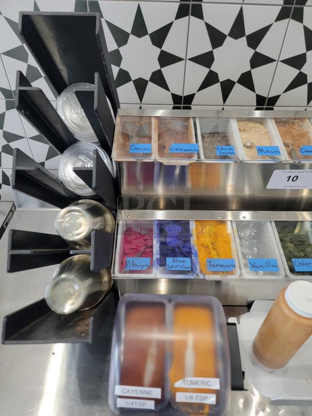 Lot of smoothie bar toppings and add-ins dispenser with labeled compartments for cacao, cinnamon, chia seeds, maca, pitaya, blue spirulina, turmeric, and more. Includes accessories and spice jars labeled cayenne and turmeric. In good condition.