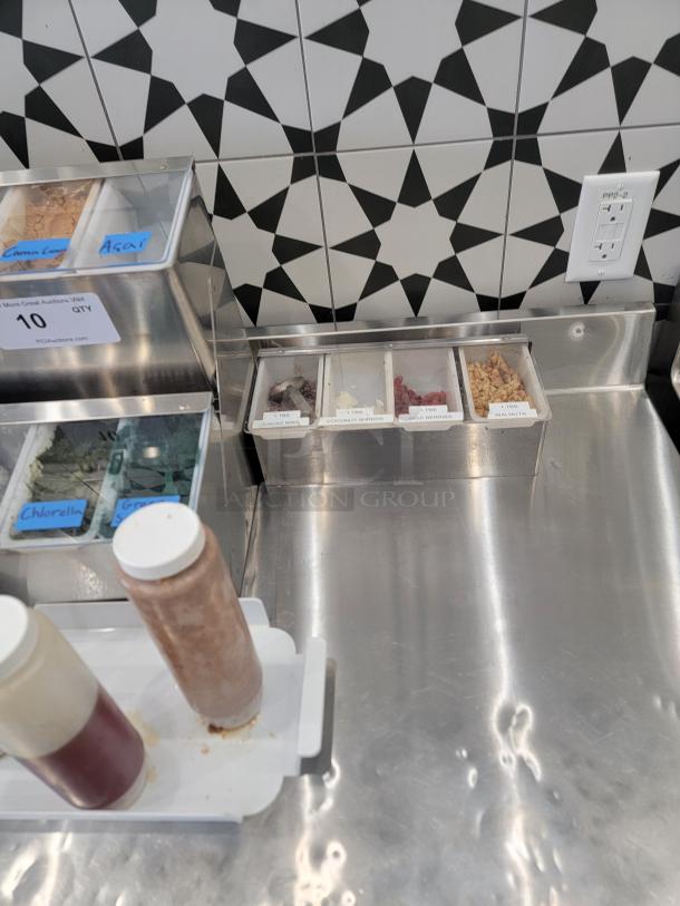 Stainless steel smoothie bar toppings dispenser with labeled compartments for cacao nibs, coconut shreds, goji berries, walnuts.