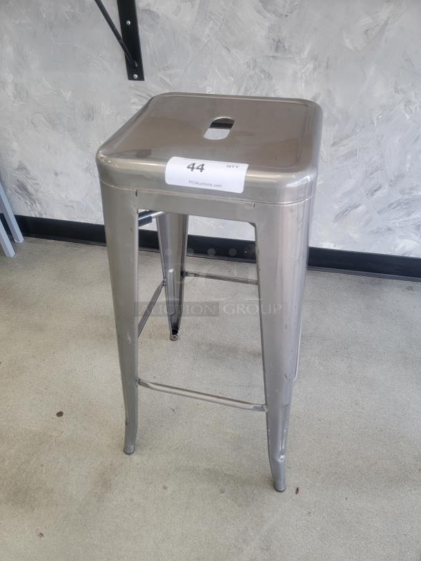 Modern industrial metal bar stool in silver/gray, stackable model, durable construction, slight surface wear, auction tag visible.