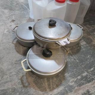 Four aluminum sauce pans with matching lids and side handles, stacked. Includes visible auction tag "4XBID." Well-used condition.