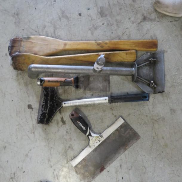 Assorted utensils on a concrete floor, including wooden paddles, metal scrapers, and a hand chopper, some showing wear.