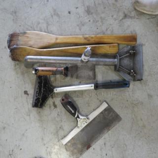 Assorted utensils on a concrete floor, including wooden paddles, metal scrapers, and a hand chopper, some showing wear.