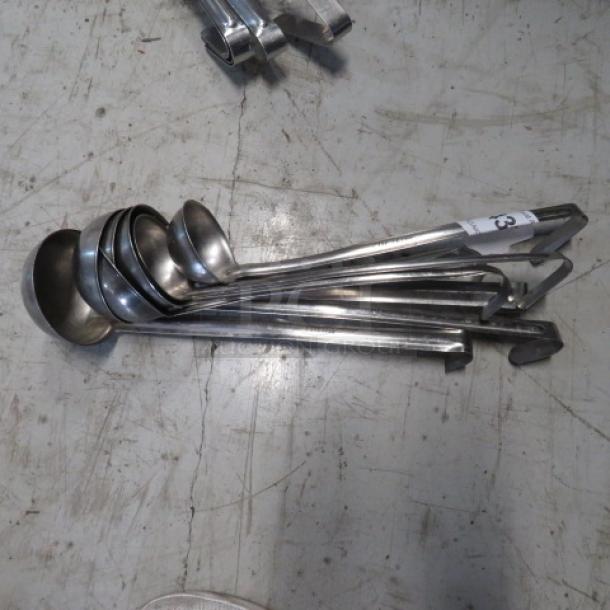Assorted stainless steel ladles in various sizes, used condition, laid out on the floor. Ladles show standard wear.