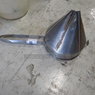Stainless steel cone strainer with a perforated design, metal handle, and visible label on a workshop surface.