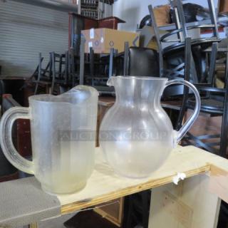 Two clear poly plastic pitchers, one fluted and one rounded, on a shelf. Used condition, with visible wear.