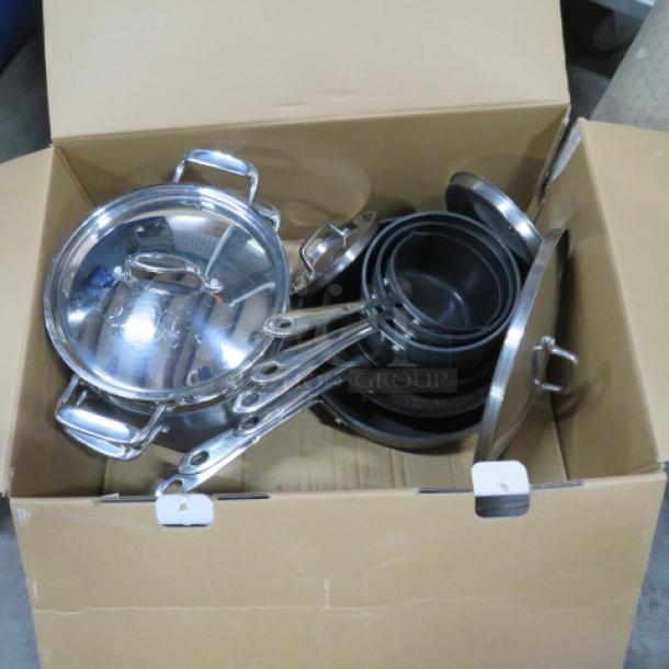 Set of stainless steel cookware in a cardboard box, includes pots, pans, lids, and handles; appears new and unused.