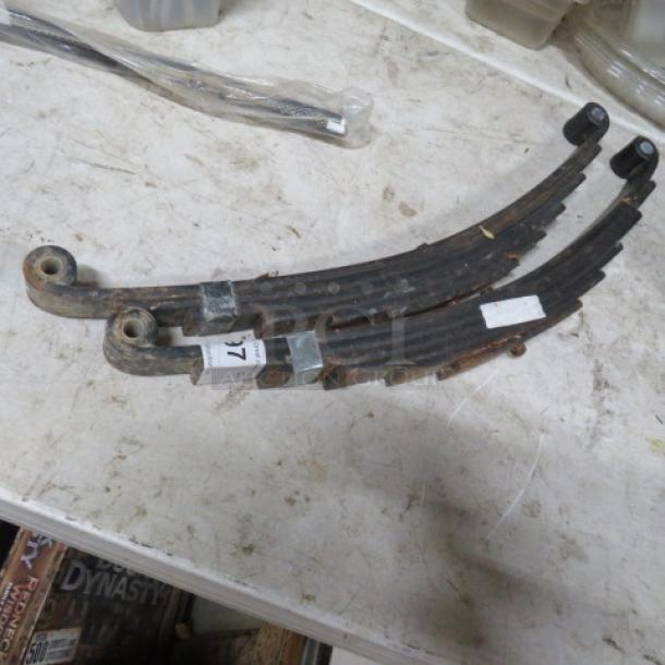 Two rusted leaf springs on a workbench, labeled as one lot. Each spring shows wear, with metal and rubber components visible.