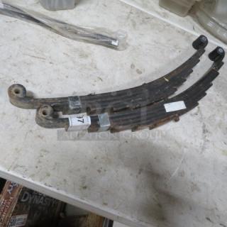 Two rusted leaf springs on a workbench, labeled as one lot. Each spring shows wear, with metal and rubber components visible.
