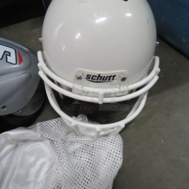 A white Schutt football helmet with faceguard, gray shoulder pads, and a white mesh jersey laid on the ground.