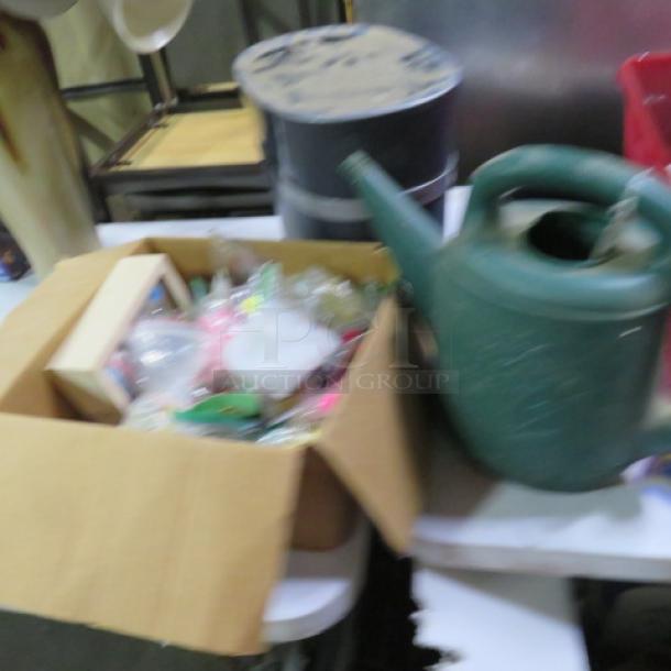 Cardboard box with assorted items and a green watering can on a table. Miscellaneous lot in used condition.