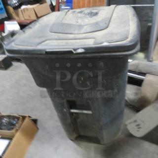 Black wheeled trash can with hinged lid, moderate wear and scratches, metal axle visible. Sturdy design for easy mobility.