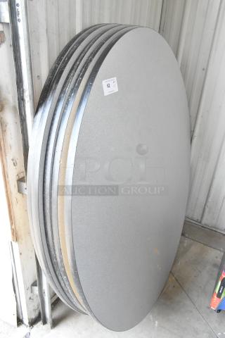 Five round gray table tops stacked against a wall. Tags indicate item 62, lot of five. Metal edging and smooth surface.