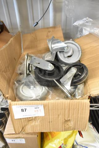 Lot of 7 commercial casters in a cardboard box. Includes various sizes and types with metal brackets. Item labeled as lot 87.