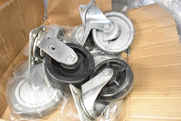 Lot of 7 commercial casters in a cardboard box. Various sizes and types, featuring metal brackets and rubber wheels. New condition.