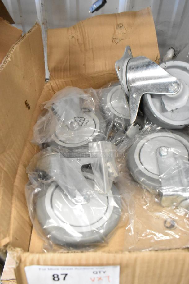 Lot of 7 commercial casters in plastic bags inside a cardboard box. Casters have metal mounts and appear unused.