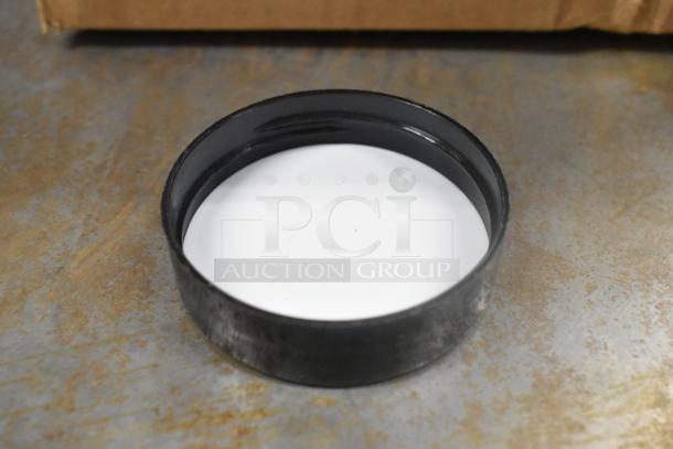 Box of black circular plastic lids, shown on industrial surface. Lids appear new, no visible markings or labels.