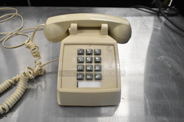 Beige corded telephone with rotary keypad, coiled cord, and standard design, positioned on a metallic surface. Vintage model.
