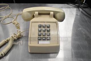 Beige corded telephone with rotary keypad, coiled cord, and standard design, positioned on a metallic surface. Vintage model.