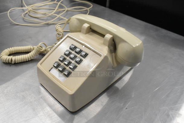 Beige corded telephone with rotary dial, coiled cable, and number pad on metal surface; vintage condition.