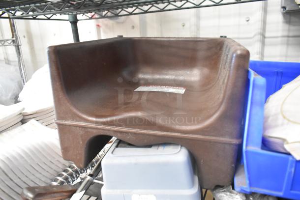 Brown booster seat with ergonomic design, visible label, and accompanying gray spray nozzle attachment underneath.