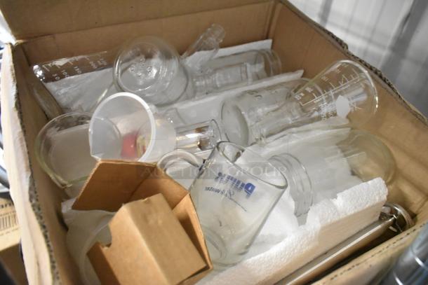 Cardboard box with various glass beakers and lab equipment, featuring visible measurement markings and labels.