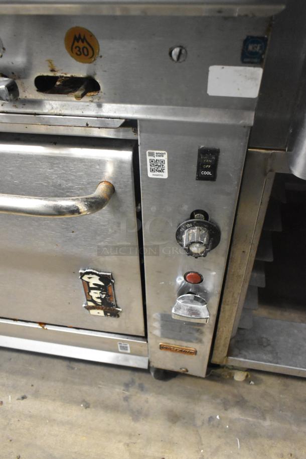 Montague stainless steel commercial gas range with 4 burners and oven. Features control knobs, NSF certified. Mounted on casters. Signs of use.