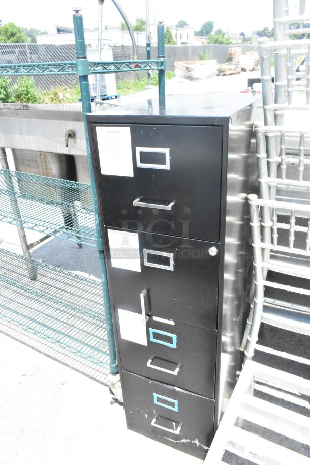 Black metal 4-drawer filing cabinet with label holders and key lock on one drawer, showing some wear.