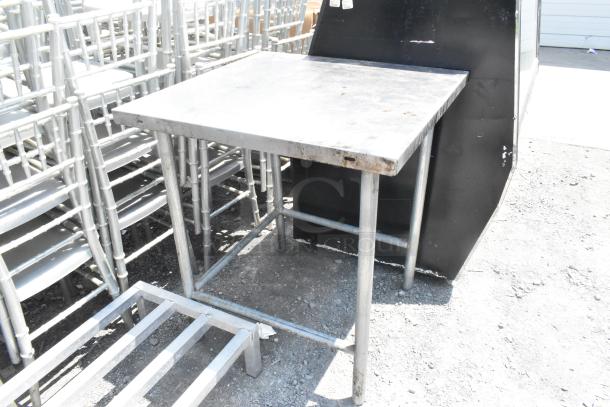 Stainless steel table with square top, visible wear, sturdy tubular legs. Suitable for industrial or kitchen use.