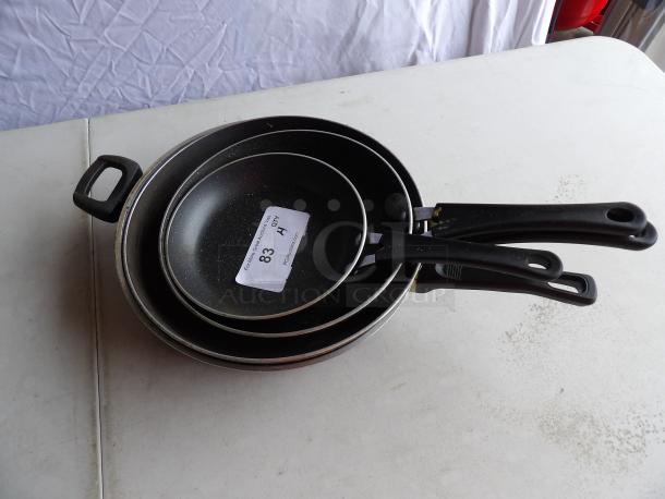 Assorted Traymontina cooking skillets, set of 4, featuring black non-stick surface and ergonomic handles. Visible label "83 QTY."