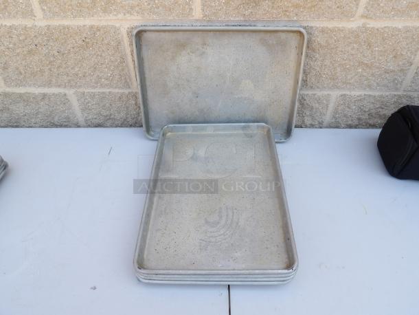 Four used half-size sheet pans with visible wear, ideal for baking. Stacked on a tabletop against a brick wall.