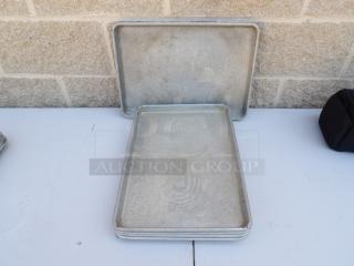 Four used half-size sheet pans with visible wear, ideal for baking. Stacked on a tabletop against a brick wall.