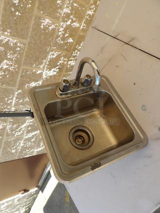Stainless steel drop-in sink with dual-handle faucet and water lines. Visible wear on edge. Suitable for kitchen use.