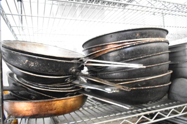 Stack of used black skillets on metal shelf; some wear visible. Part of a lot with stainless steel bins and metal bowls.