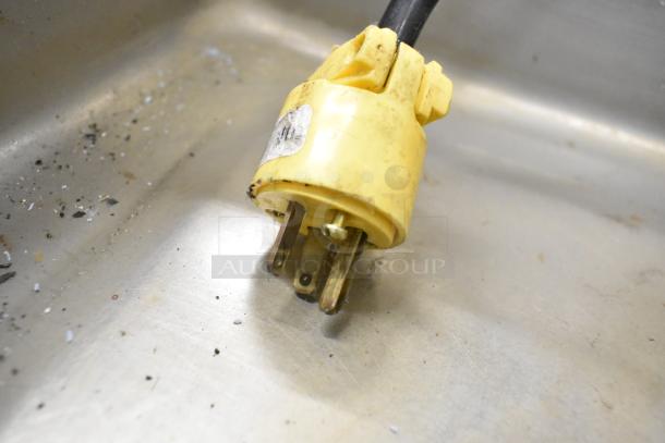 A yellow electrical plug with metal prongs lies on a stainless steel surface, showing signs of wear.