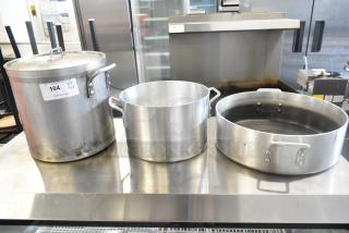 Three metal stock pots on a stainless steel surface, varying in size, with handles. Visible wear. Auction tag "104, Qty x3."