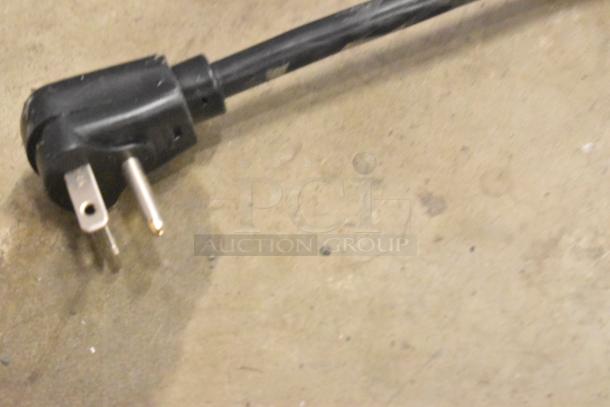 A close-up of a black, slightly angled electrical plug with three prongs on a concrete floor.