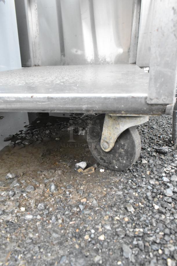 Metal 3 tier cart with commercial casters, visible one caster, slightly worn, on gravel surface.
