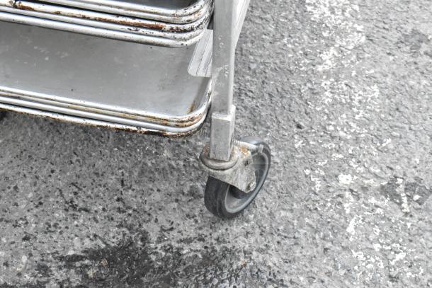 Metal commercial pan transport rack on caster with 13 full-size metal baking pans, some wear visible on frame.