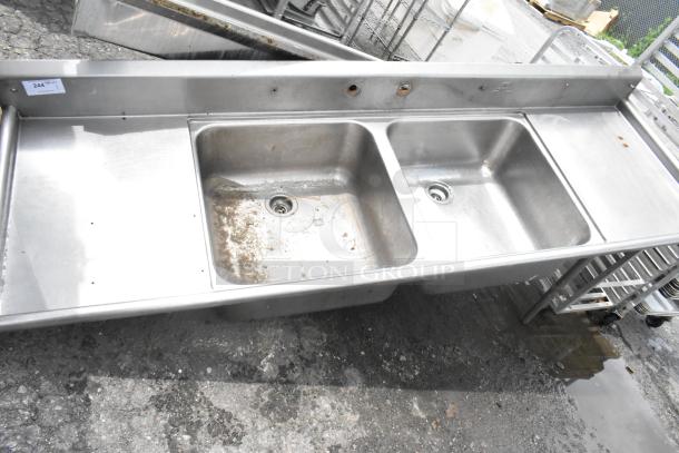 Stainless steel 2-bay sink with dual drain boards, no legs. Features two faucet holes and some surface wear, ideal for commercial use.