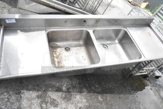Stainless steel 2-bay sink with dual drain boards, no legs. Features two faucet holes and some surface wear, ideal for commercial use.