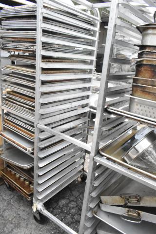 Metal pan transport rack with multiple shelves on commercial casters, holding various baking sheets and pans, in used condition.