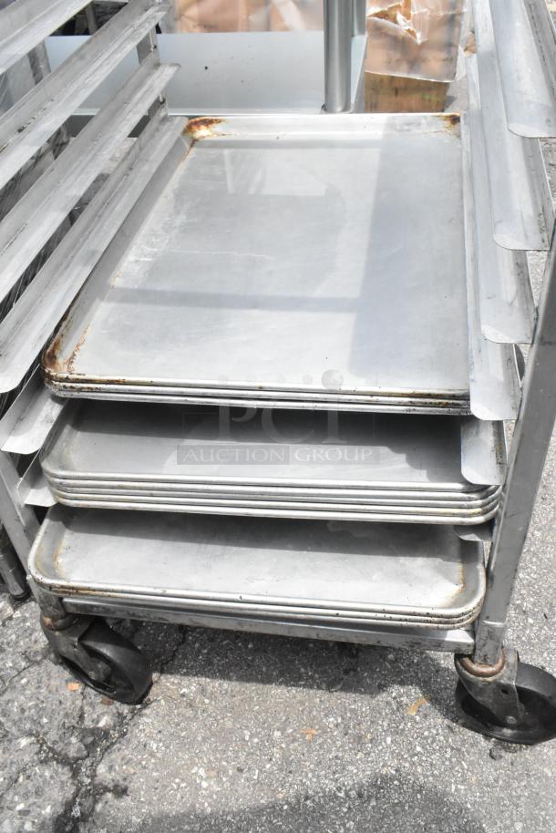 Metal pan transport rack with 11 metal baking pans on commercial casters, showing wear. Ideal for bakery use.