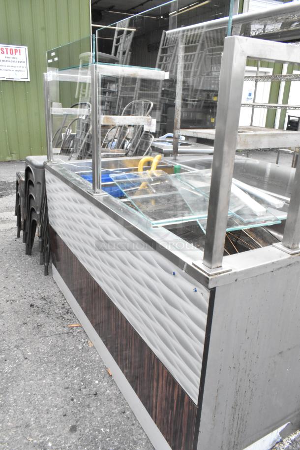 Metal commercial buffet station with sneeze guard, featuring a glass top and textured metal and wood paneling. Good condition.