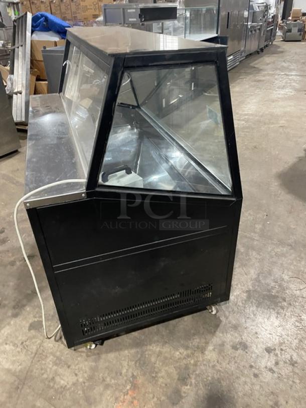 Commercial black slanted glass refrigerated bakery/deli display case with rear prep area, lower storage, and sliding doors on casters.