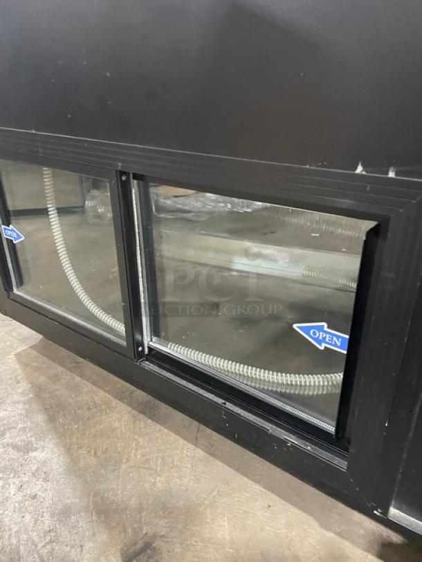 Commercial black slanted glass refrigerated bakery display case with rear prep area, lower storage, and sliding doors on casters.