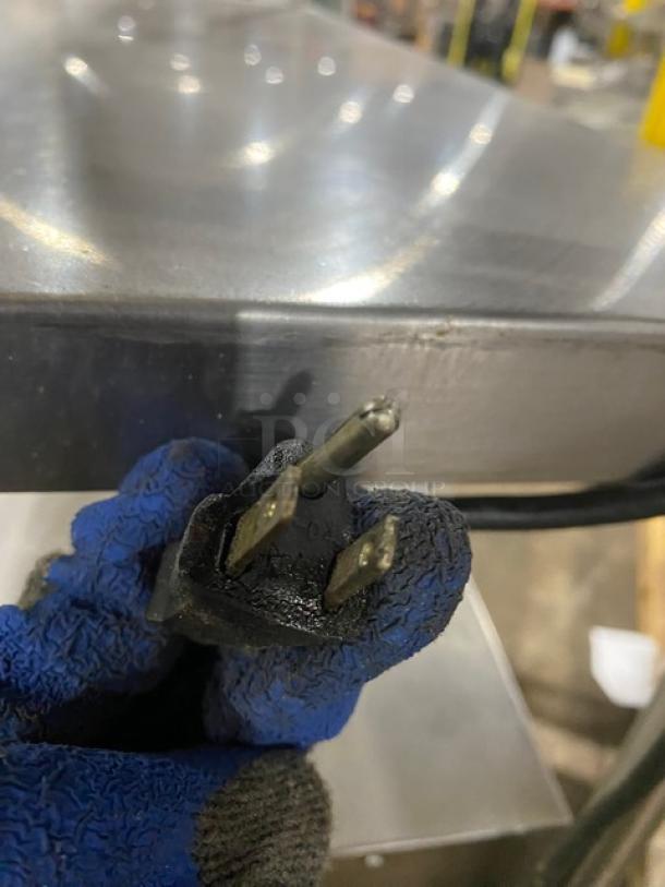 Close-up of an electrical plug held by a gloved hand, with a stainless steel surface in the background.
