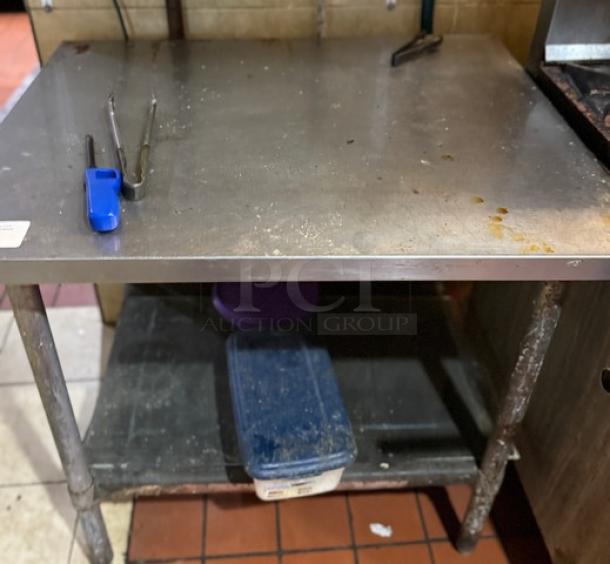 Stainless steel work table with tubular legs, used condition, shows stains and wear, includes lower storage shelf.