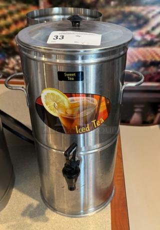 Stainless steel tea/beverage dispenser with lid, tap, and handles. Labels indicate "Sweet Tea" and "Iced Tea." Auction tag visible.